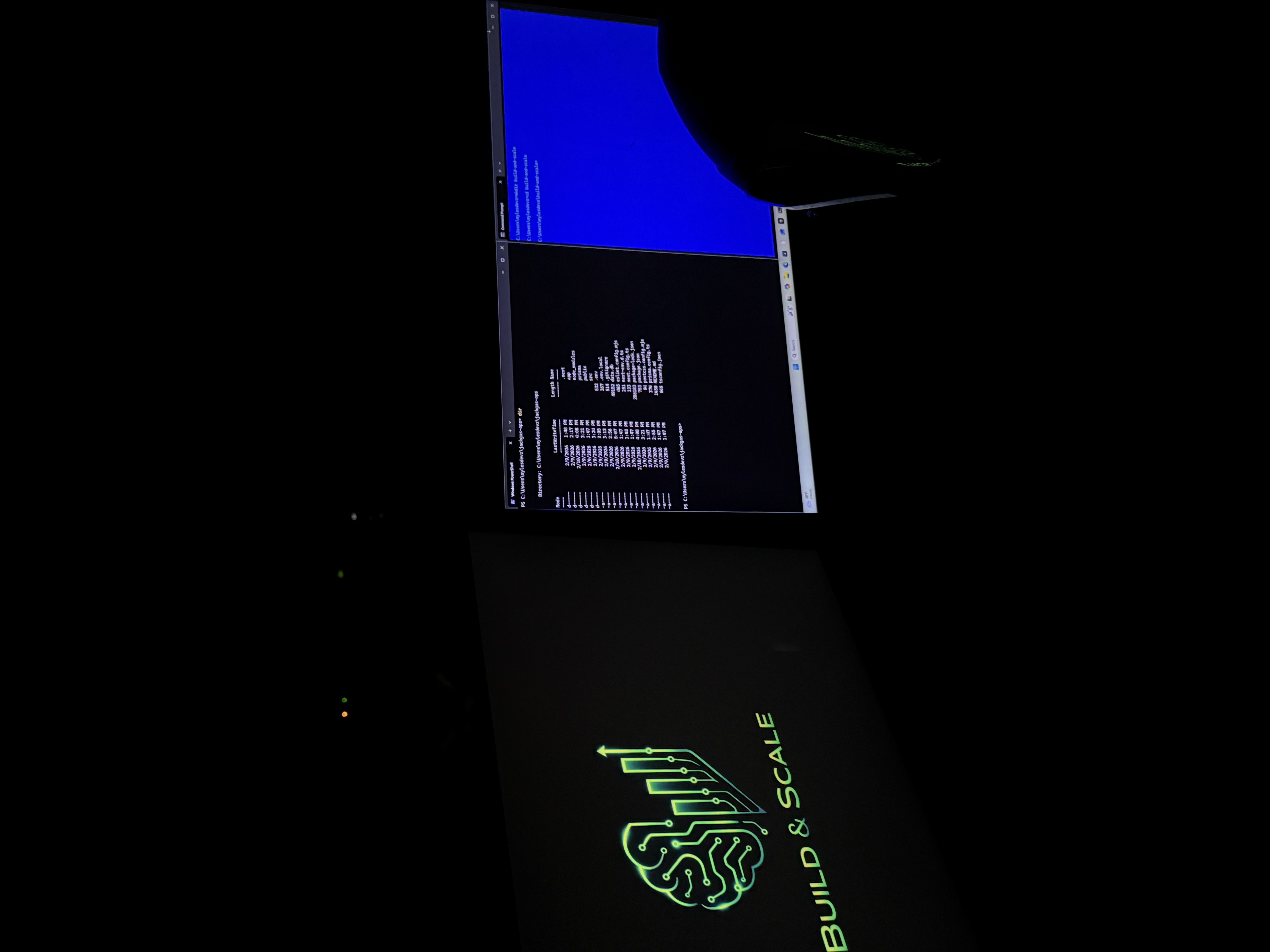 Build & Scale workspace: dual monitors on wooden desk, left screen showing company logo with circuit-brain and growth chart, right screen with terminal. Teal and blue glow.